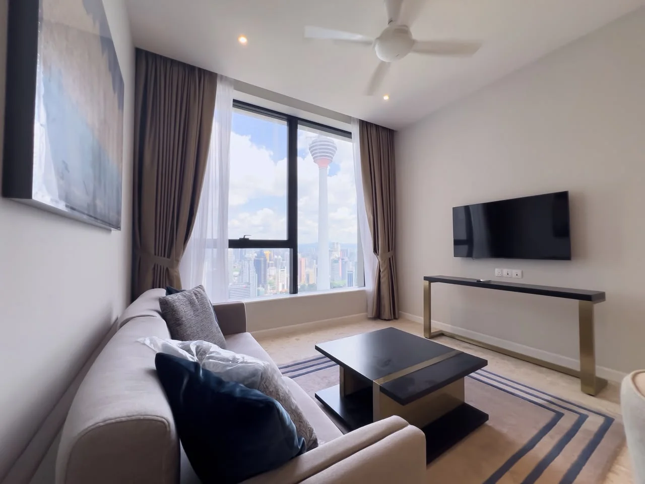 Living features: 3-seater fabric sofa in light grey, decorative cushions in blue, glass coffee table with gold legs, wall-mounted flat-screen TV, floor-to-ceiling windows, curtains in light brown, recessed downlights, carpet with geometric pattern, city skyline view, ceiling fan; style: modern; view: city.